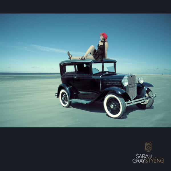 1920s car in black. lady dressed in 1920s black flapper dress with red hat, sitting on roof of car